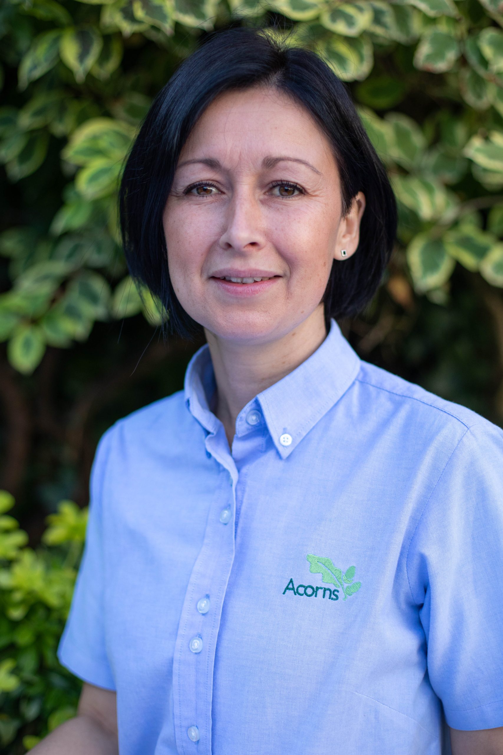 Acorns Nurseries A person with short dark hair in a light blue shirt featuring an Acorns logo stands serenely amid the lush green foliage, evoking the tranquil charm of Llandaff.