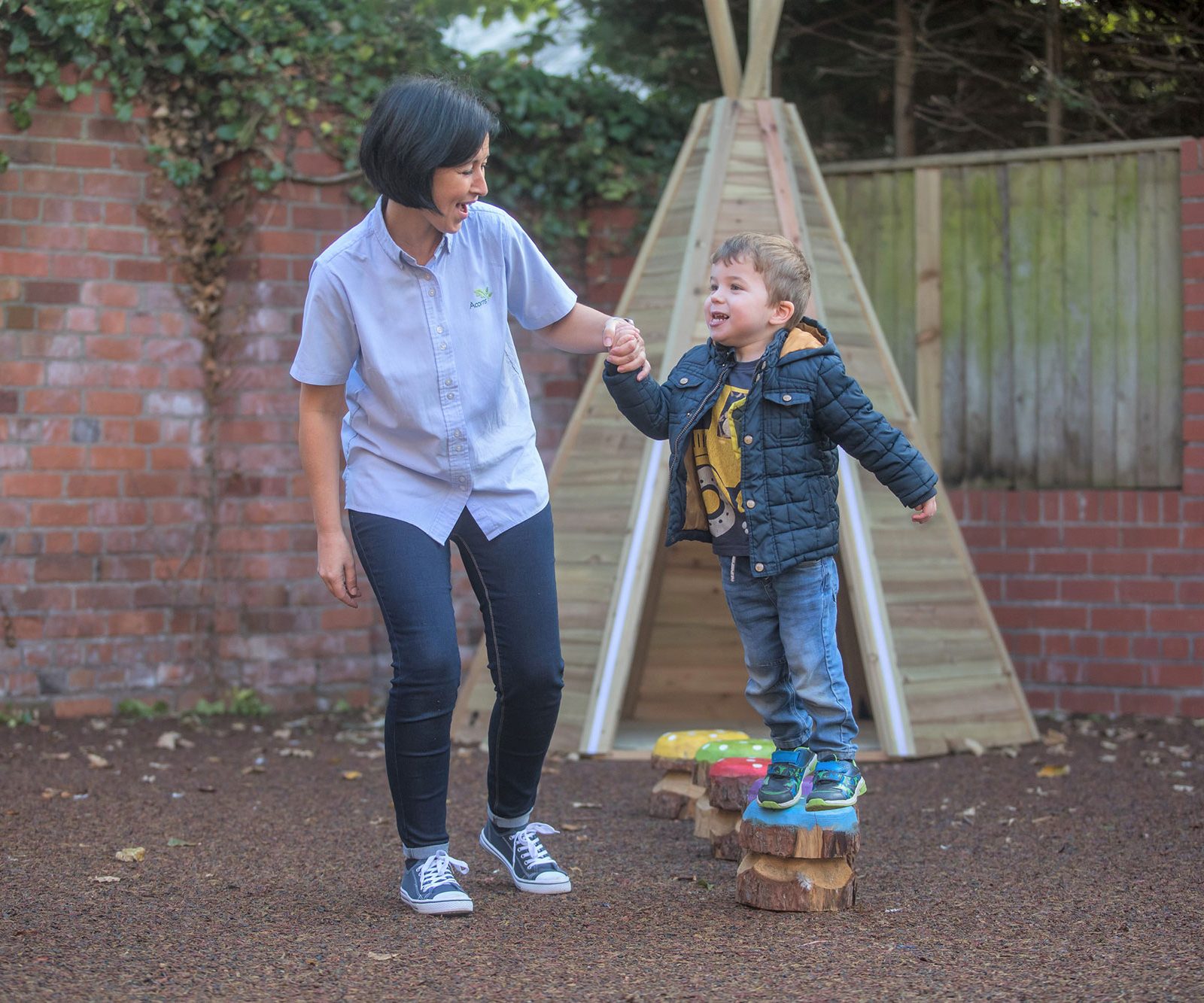 Acorns Nurseries An adult gently holds a child's hand as the little one balances on wooden stepping stones in Llandaff's charming outdoor playground, with an Acorns-inspired teepee standing proudly in the background.