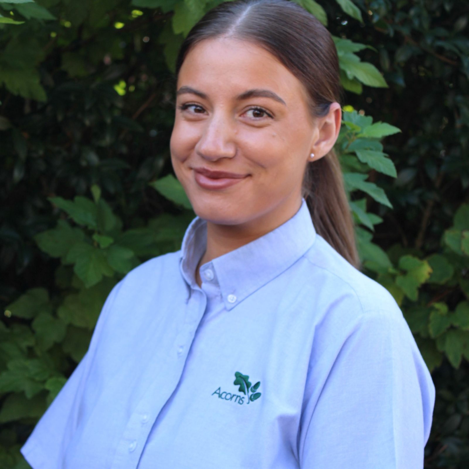 Acorns Nurseries A woman in a light blue shirt with an embroidered logo stands smiling against a leafy backdrop, exuding the natural charm of Llandaff.
