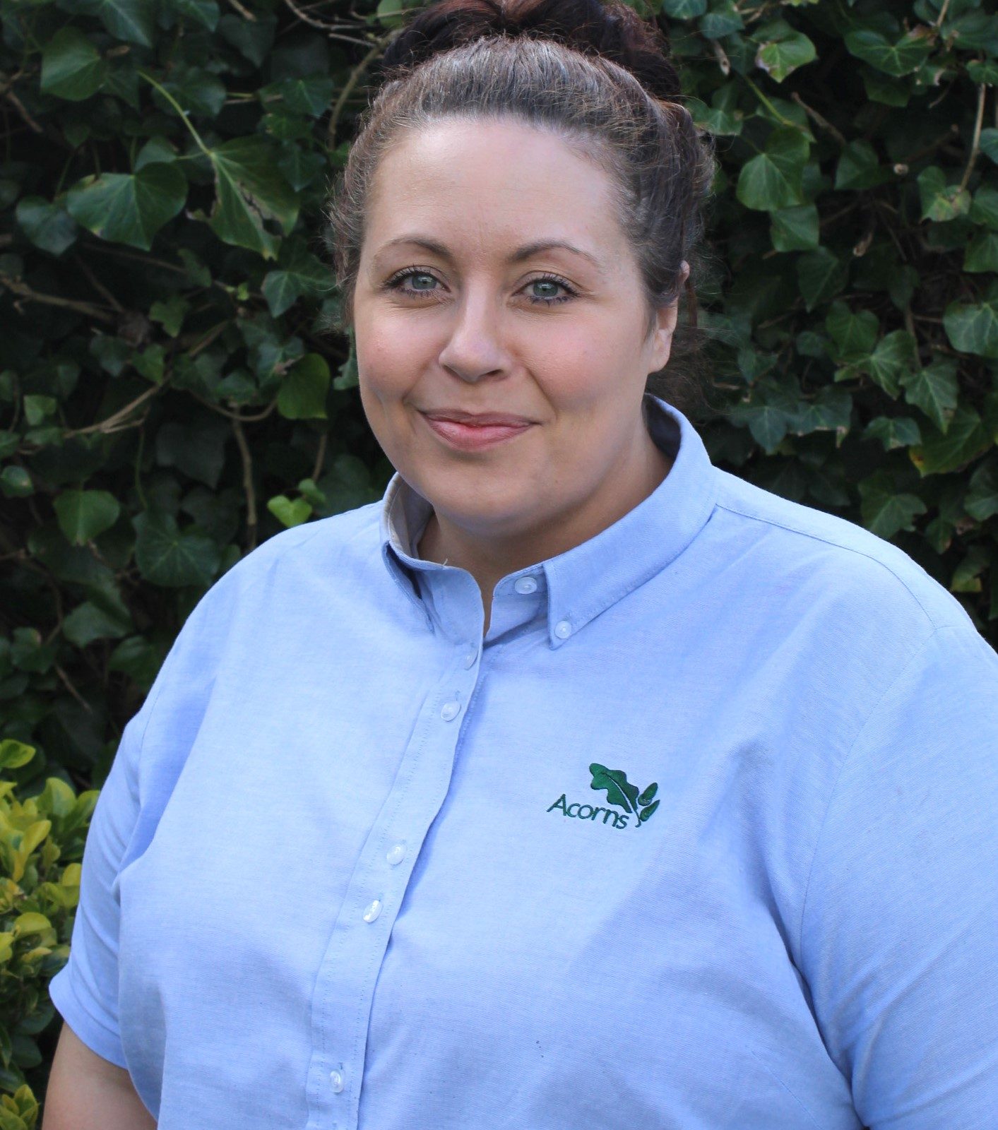 Acorns Nurseries Woman in a light blue Acorns House shirt, smiling slightly at the camera against a leafy backdrop.