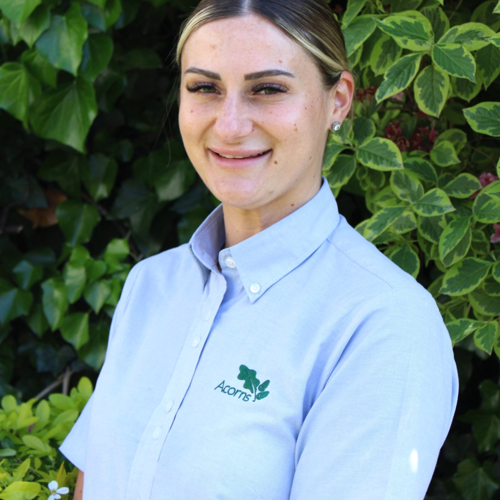Acorns Nurseries Woman in a light blue Acorns shirt smiles by green bushes outside at Hill House.