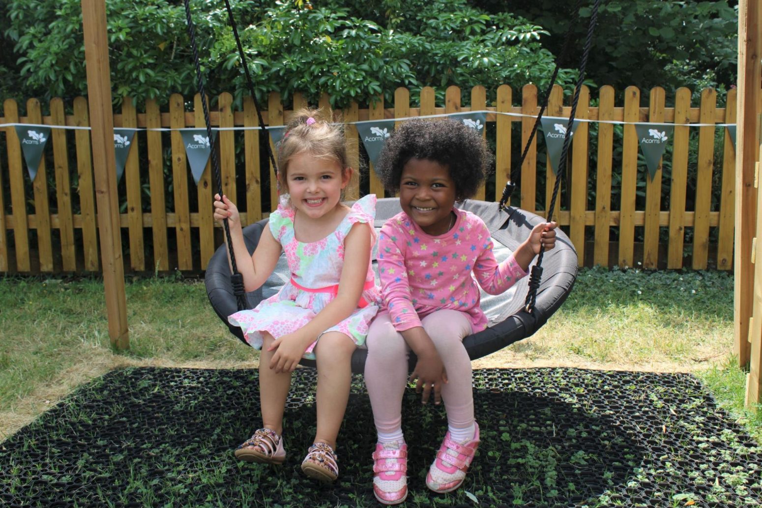 Acorns Nurseries Two young girls, carefree and laughing, sit together on a swing in a garden dotted with acorns, with a wooden fence and towering trees in the background. Acorns Nurseries Two young girls, carefree and laughing, sit together on a swing in a garden dotted with acorns, with a wooden fence and towering trees in the background.