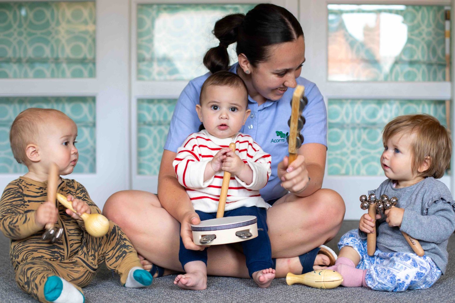 Acorns Nurseries A woman sits on the floor with three infants, each holding a percussion instrument, in a brightly lit room decorated with acorns. Acorns Nurseries A woman sits on the floor with three infants, each holding a percussion instrument, in a brightly lit room decorated with acorns.