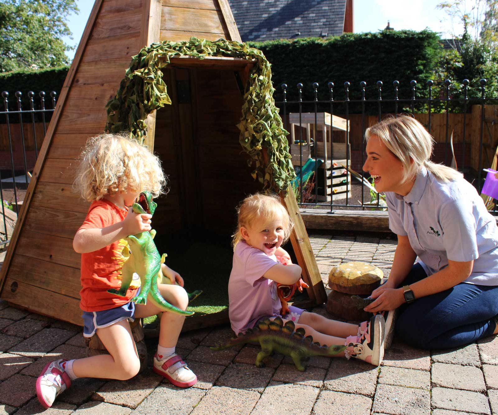Acorns Nurseries Two young children play with toy dinosaurs beside a wooden play structure in Llandaff, while a woman kneels nearby, smiling and engaging with them. Acorns scatter across the ground, adding to the scene's charm.