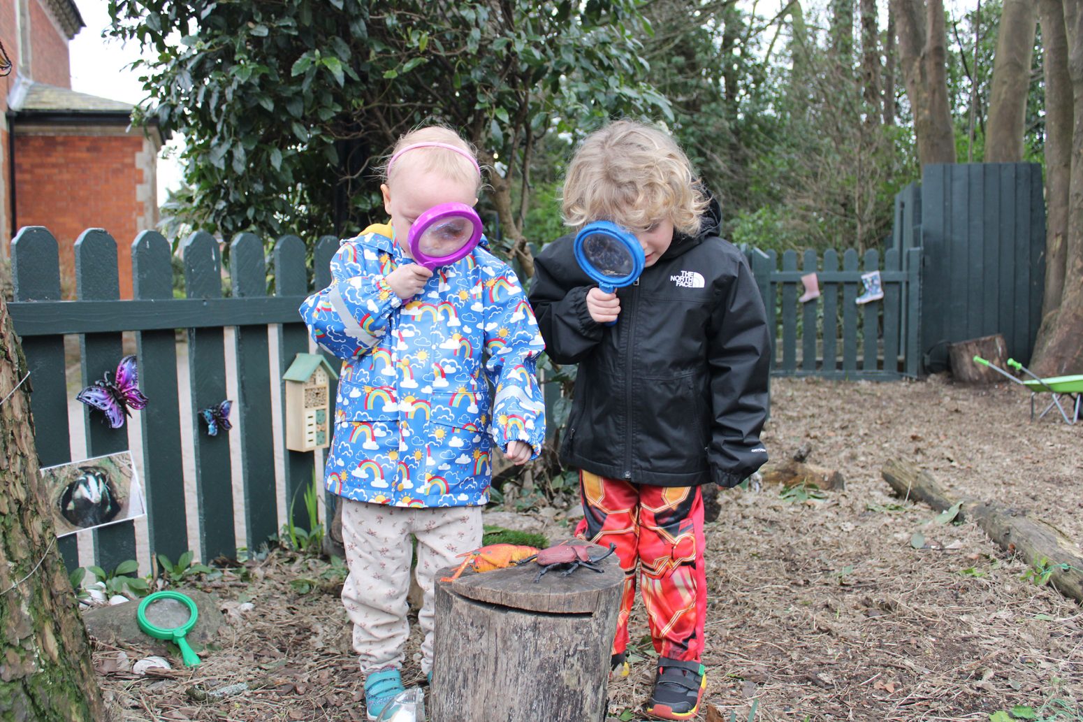 Acorns Nurseries Two children in colorful jackets examine a tree stump with magnifying glasses, discovering acorns nestled in its crevices, in a lively garden setting. Acorns Nurseries Two children in colorful jackets examine a tree stump with magnifying glasses, discovering acorns nestled in its crevices, in a lively garden setting.