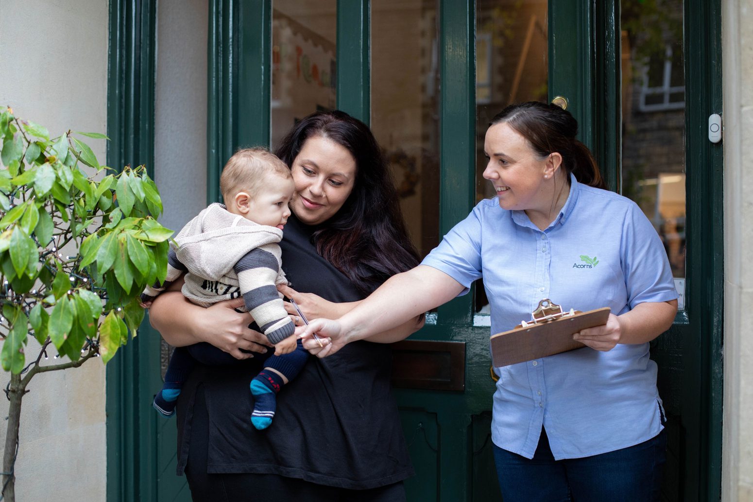 Acorns Nurseries A woman holding a baby interacts with another woman registering details on a clipboard in front of a doorway adorned with a small potted acorn plant. Acorns Nurseries A woman holding a baby interacts with another woman registering details on a clipboard in front of a doorway adorned with a small potted acorn plant.