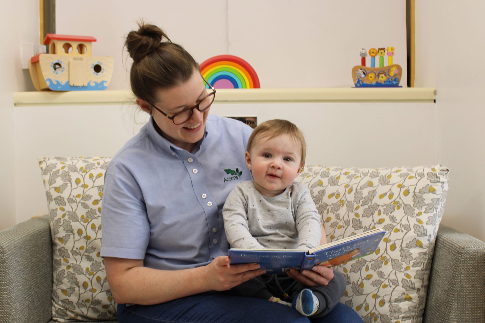 Acorns Nurseries A person with glasses reads a book to a young child on a couch, surrounded by colorful toys and Acorns on the shelf behind them. Acorns Nurseries A person with glasses reads a book to a young child on a couch, surrounded by colorful toys and Acorns on the shelf behind them.