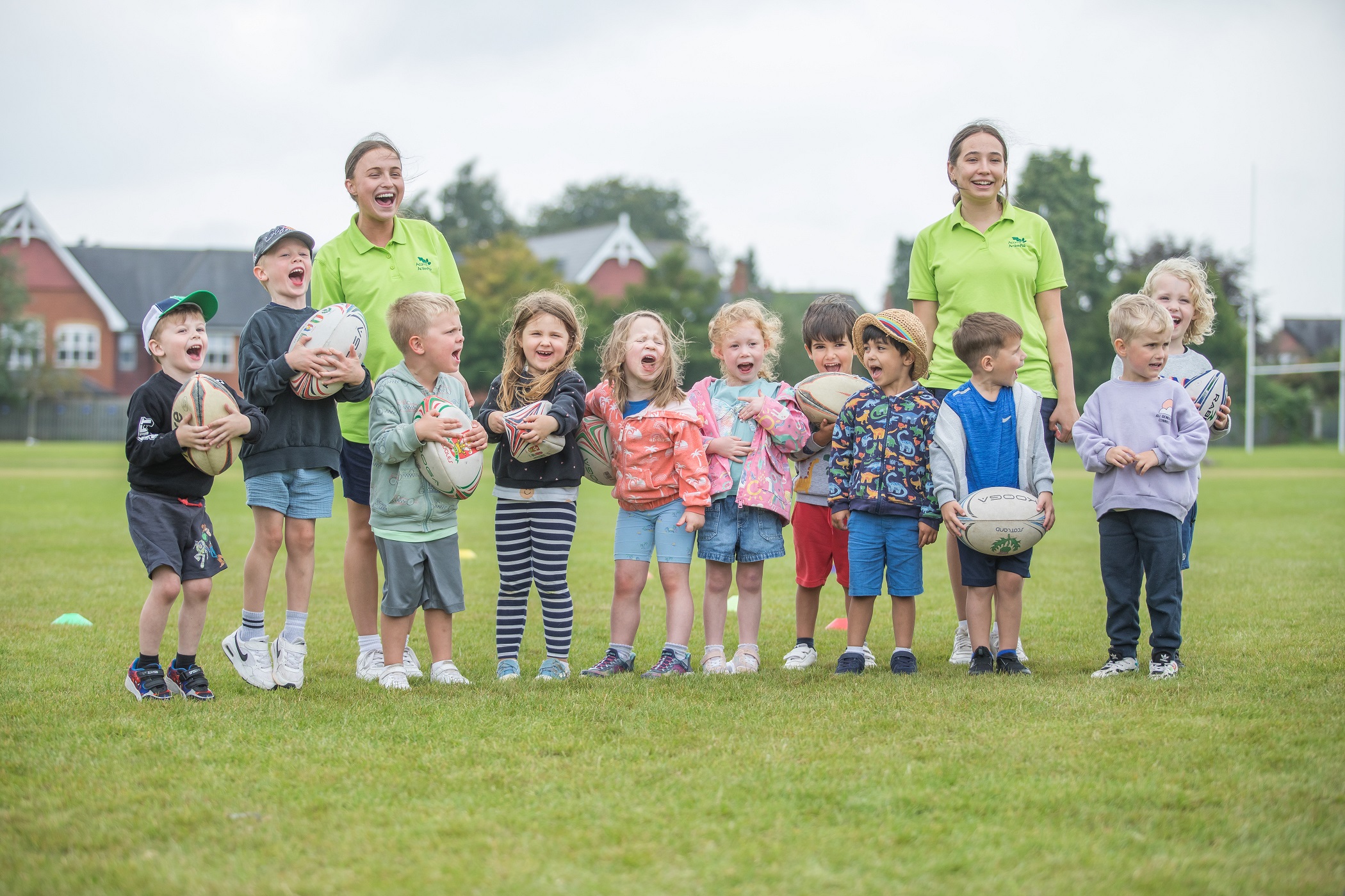 Acorns Nurseries A group of children and two adults stand on grass, smiling with rugby balls at Action Pak, Summer 2025.