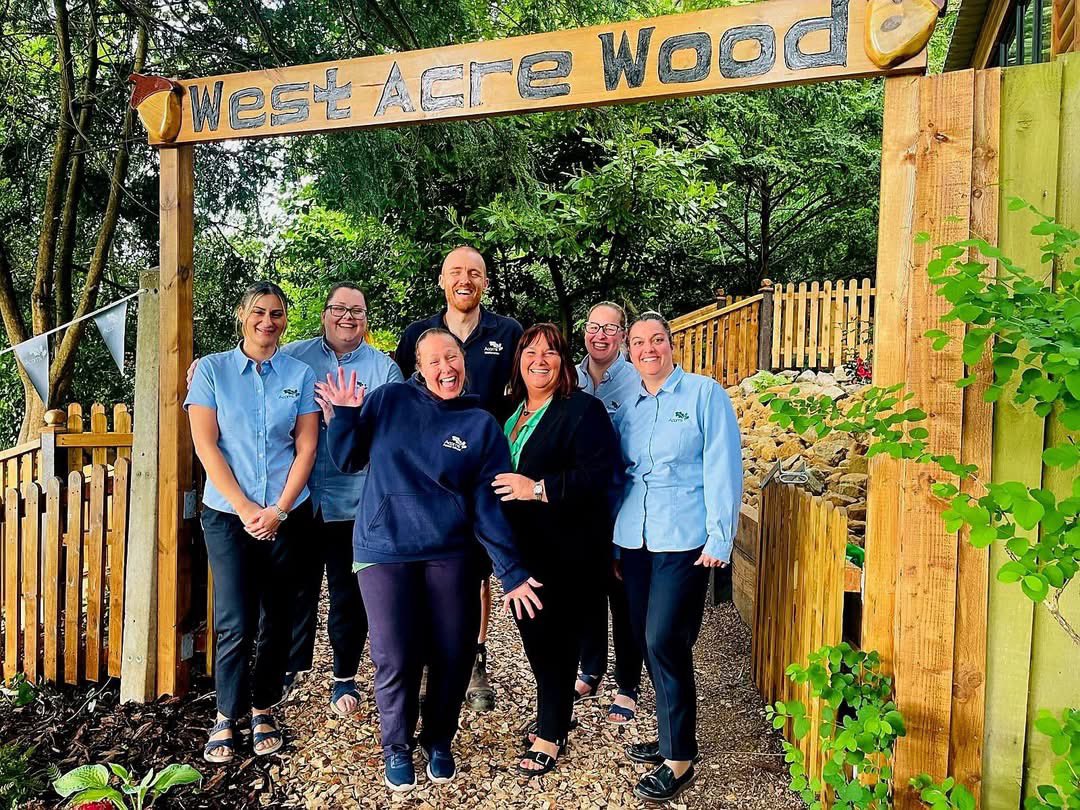 Acorns Nurseries Six smiling adults stand under a "West Acre Wood" sign at Forest School, surrounded by trees and wooden fencing.
