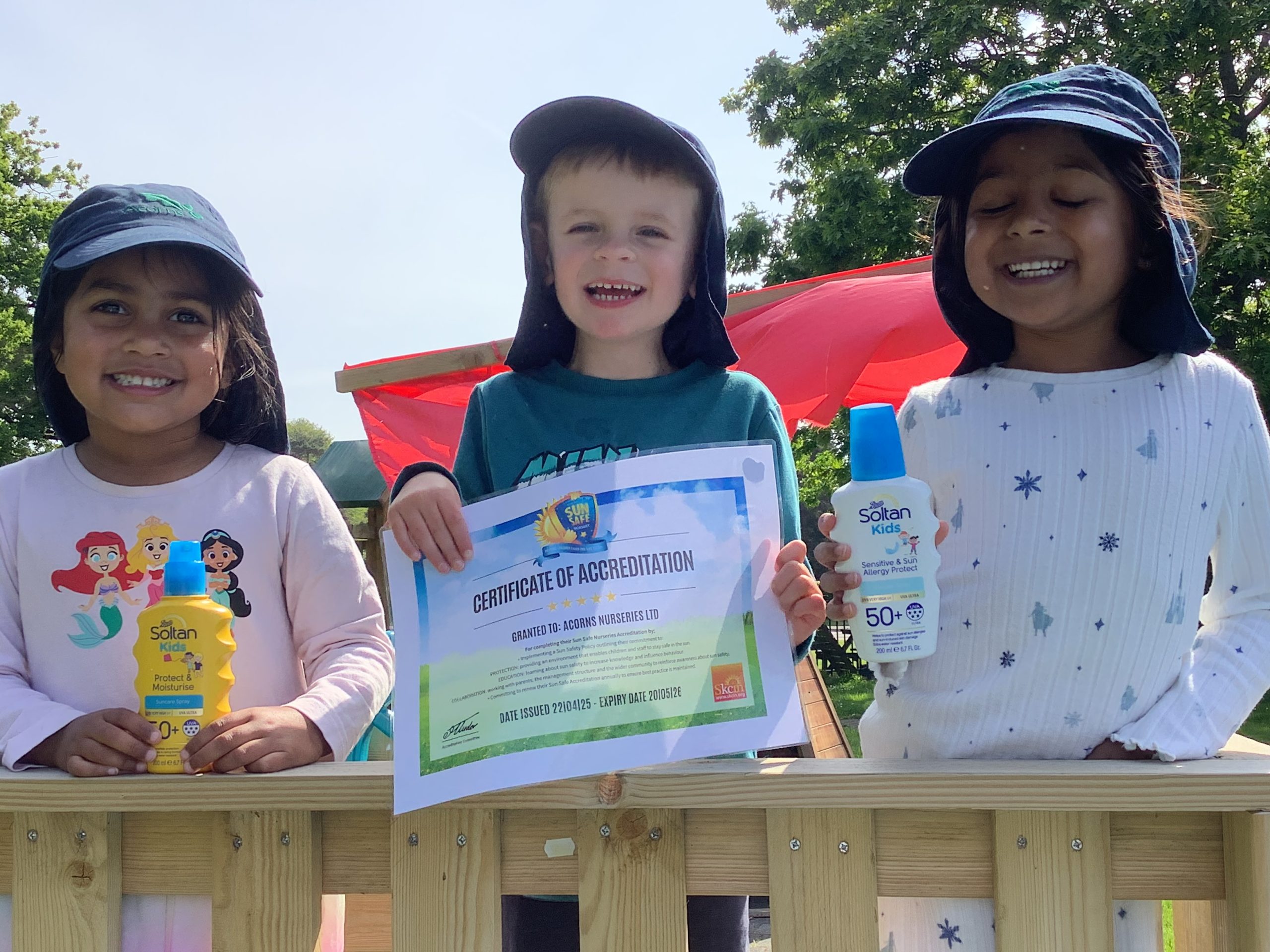 Acorns Nurseries Three smiling children in sun hats hold sunscreen and a Sun Safe Accreditation certificate by a wooden fence outdoors.